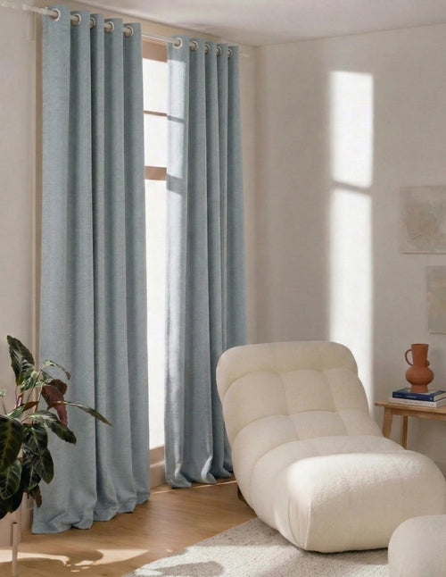Rideau sur mesure bleu ciel Kurtens à œillets dans salon baigné de lumière avec fauteuil design crème et plante tropicale